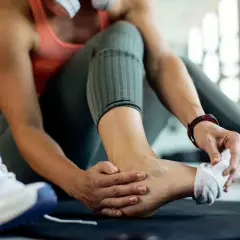 Kobieta z bólem kostki, trzymająca się za stopę, obok butów sportowych.