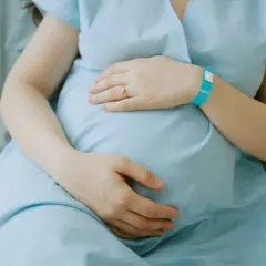 Ciężarna kobieta w niebieskiej koszuli szpitalnej trzymająca się za brzuch, z opaską na nadgarstku.