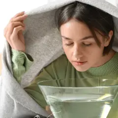 Kobieta z zamkniętymi oczami pochyla się nad miską z wodą, przykryta kocem, wykonując inhalację.