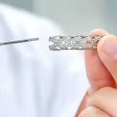 Dłoń trzyma metalowy stent o ażurowej strukturze, obok widoczny jest instrument medyczny.