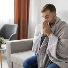 Mężczyzna owinięty kocem siedzi na kanapie i wyciera nos chusteczką.