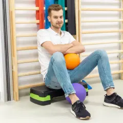 Fizjoterapeuta siedzi na schodku z piłkami, na tle drabinek i sprzętu rehabilitacyjnego.
