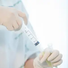 Ręce w rękawiczkach trzymają strzykawkę z płynem i probówkę.