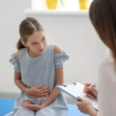 Dziewczynka w niebieskiej sukience trzyma się za brzuch podczas wizyty u lekarza.