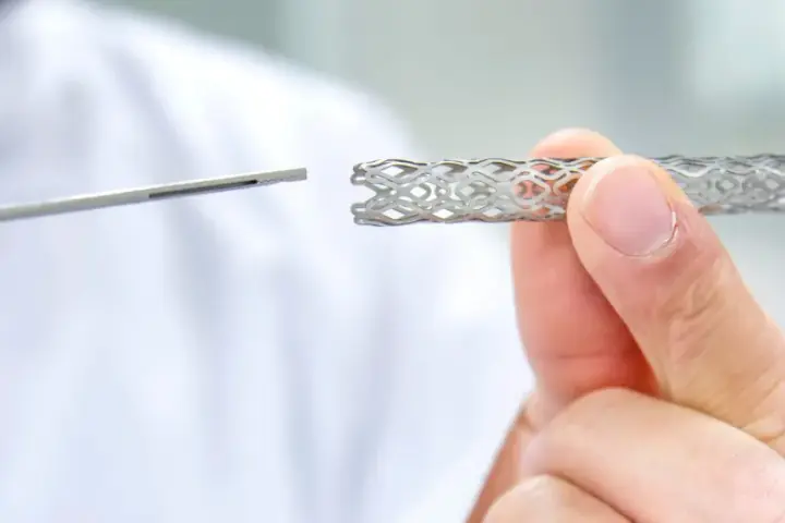 Dłoń trzyma metalowy stent o ażurowej strukturze, obok widoczny jest instrument medyczny.