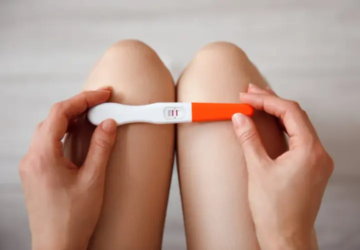 Dłonie trzymające test ciążowy z dwoma kreskami, na tle nóg.