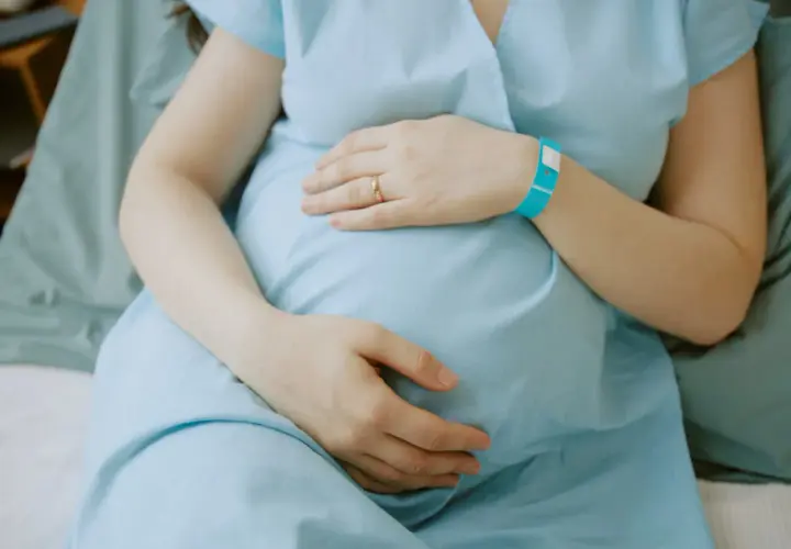 Ciężarna kobieta w niebieskiej koszuli szpitalnej trzymająca się za brzuch, z opaską na nadgarstku.