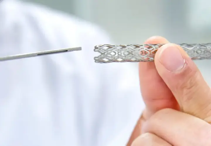 Dłoń trzyma metalowy stent o ażurowej strukturze, obok widoczny jest instrument medyczny.