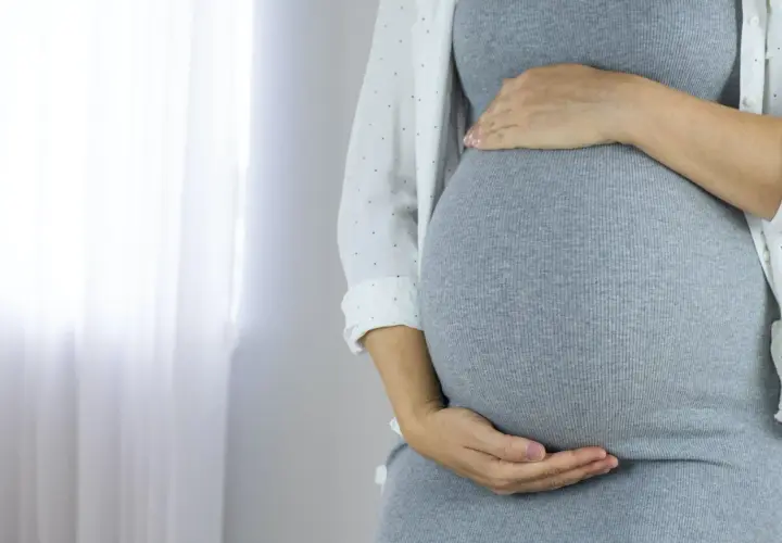 Zbliżenie na brzuch ciężarnej kobiety w szarej sukience, trzymający się za brzuch.