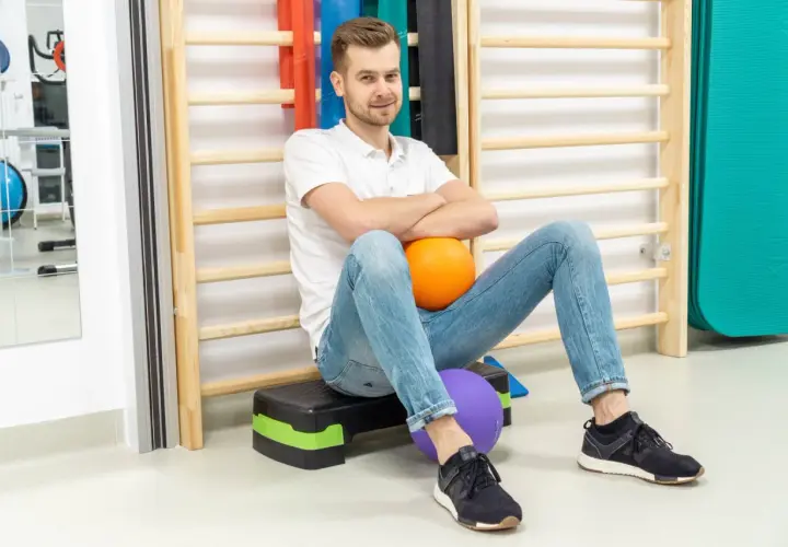 Fizjoterapeuta siedzi na schodku z piłkami, na tle drabinek i sprzętu rehabilitacyjnego.