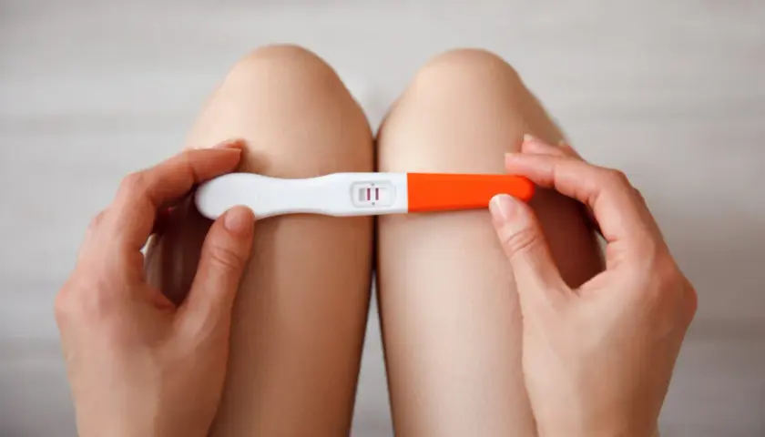 Dłonie trzymające test ciążowy z dwoma kreskami, na tle nóg.