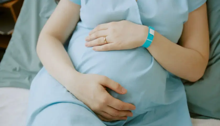 Ciężarna kobieta w niebieskiej koszuli szpitalnej trzymająca się za brzuch, z opaską na nadgarstku.