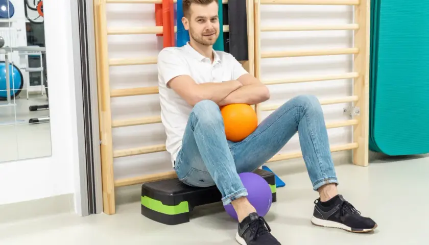 Fizjoterapeuta siedzi na schodku z piłkami, na tle drabinek i sprzętu rehabilitacyjnego.