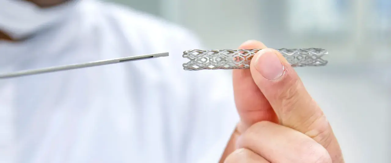 Dłoń trzyma metalowy stent o ażurowej strukturze, obok widoczny jest instrument medyczny.